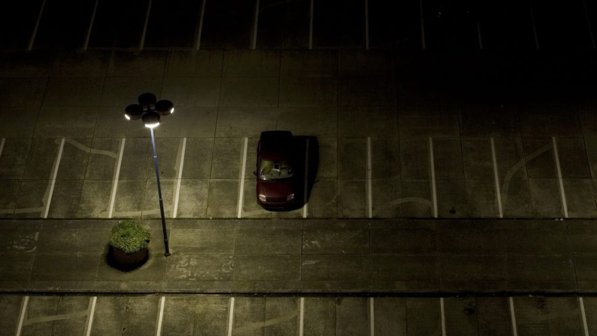 A lone car in a parking lot at night underneath a lamplight.